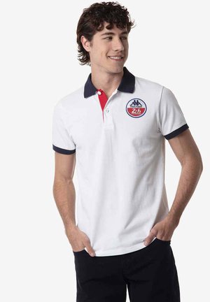 Jonge man die lacht, draagt een wit poloshirt met marineblauwe kraag en mouwranden, rode knoopsluiting en een rond racelogo op de borst.