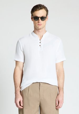 Homme portant des lunettes de soleil noires, une chemise blanche texturée à manches courtes avec boutons et un pantalon beige, debout devant un fond clair uni.
