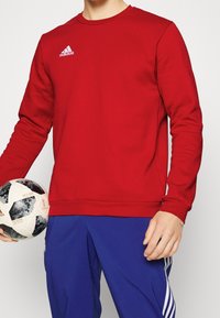 Rood crewneck sweatshirt van zacht materiaal, met een wit Adidas-logo op de borst. Gecombineerd met blauwe sportbroeken en een zwart-witte voetbalsbal.