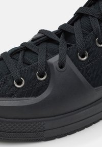 Gros plan d'une basket noire avec tissu texturé, œillets métalliques, lacets tressés épais et embout ainsi que semelle en caoutchouc protecteur.
