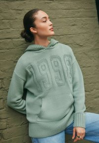 Zielona, dziergana bluza z kapturem z teksturowym wzorem "1998" na przodzie, z kangurzą kieszenią i ściągaczami na rękawach, noszona z niebieskimi dżinsami.