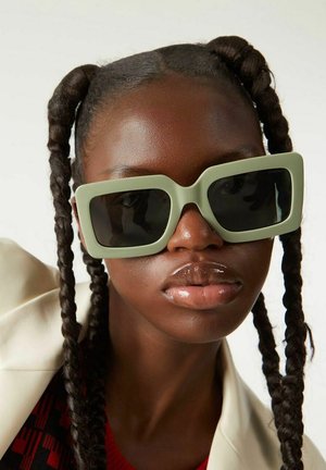 Lunettes de soleil carrées en vert discret, avec des verres foncés et des montures épaisses. Le modèle a des cheveux tressés et porte une veste de couleur claire sur un haut à motifs.