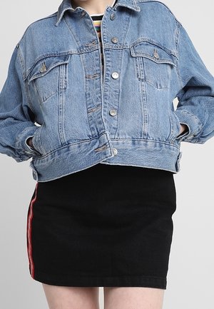 Personne portant une veste en denim bleue avec des boutons, par-dessus une jupe noire avec des rayures rouges sur le côté, les mains dans les poches de la veste.