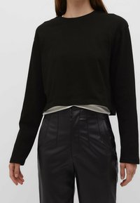 Zwarte langmouwen crop-top in een zachte stof, gelaagd over een lichtgekleurde top, gecombineerd met hoog-getailleerde zwarte nepleer broek.