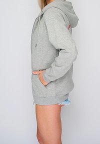 Felpa grigia con tasca frontale, cappuccio con coulisse e polsini a costine. Indossata con pantaloni corti in denim con orlo sfrangiato e dettagli azzurri.