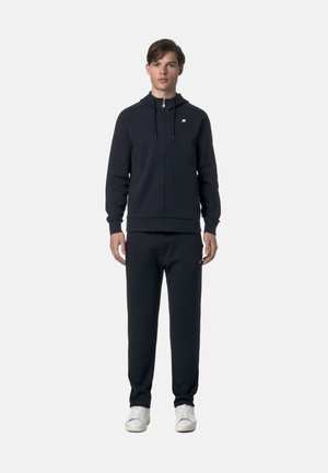 Giovane uomo in piedi che indossa un hoodie nero con zip e coulisse, pantaloni neri abbinati e scarpe da ginnastica bianche, su sfondo grigio chiaro uniforme.
