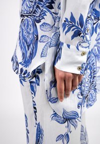 White shirt with blue botanical print, lightweight fabric, long sleeves, button closure at cuffs, rounded hem, and textured floral accents.