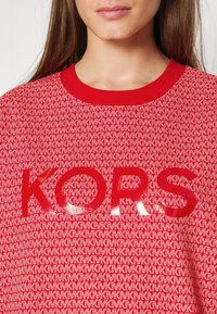 Felpa rossa in tessuto strutturato con motivo MK ripetuto e logo "KORS" in bold lucido al centro. Collo a coste.