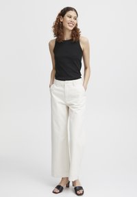 Top sans manches noir, pantalon large blanc, texture légère ; le modèle porte des sandales à talons carrés noirs, les mains dans les poches, fond neutre.