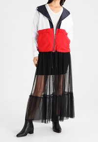 Czerwono-biały kurtka przeciwwiatrowa na zamek błyskawiczny z granatowym kapturem; zestawiona z przejrzystą czarną spódnicą z tiulu i botkami za kostkę, posiadającymi warstwową teksturę.