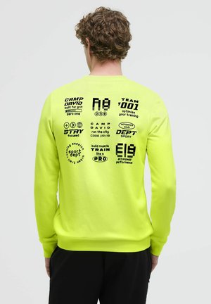 Mann mit lockigem Haar, der ein leuchtend gelbes Langarmshirt mit schwarzem, aufgedrucktem athletischem und motivierendem Text auf dem Rücken trägt.