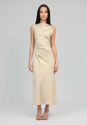 Femme aux longs cheveux bruns portant une robe midi en satin beige sans manches et des talons bruns à brides, posant devant un fond gris uni.