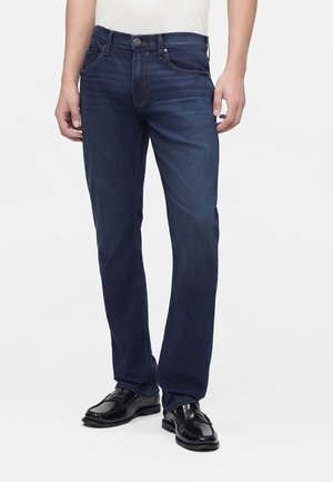 Man draagt donkerblauwe rechte jeans met zwarte leren nette schoenen en een wit overhemd, staand voor een effen achtergrond.