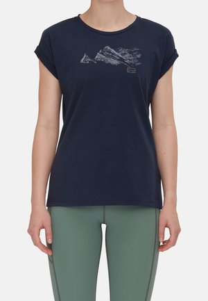 Femme portant un t-shirt bleu marine avec un graphique de montagne et des manches retroussées, associé à un legging de sport vert.