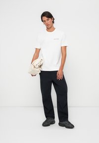 White cotton t-shirt with small black logo, paired with loose-fitting black cargo pants and dark athletic shoes, holding a beige bag.