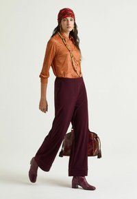 Camicia arancione a pois, pantaloni ampi color amaranto, stivaletti viola in pelle di serpente e una borsa marrone con nappine. Capelli avvolti in una sciarpa decorata.