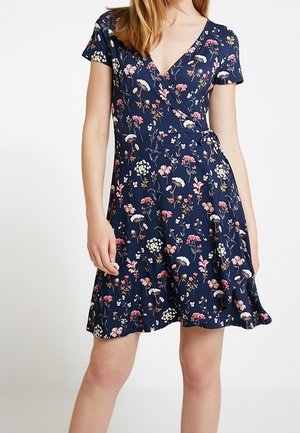 Laivastonsininen lyhythihainen kietaisumekko, jossa on vaaleanpunainen, valkoinen ja vihreä kukkakuvio, yllään vaaleaihoisella ja vaaleanruskeat laineikkaat hiukset omaavalla naisella.