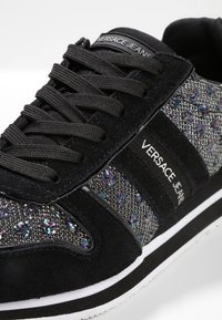 Baskets Versace Jeans présentant un mélange de daim noir et de maille métallisée, avec des accents scintillants, des lacets noirs, et une semelle blanche.