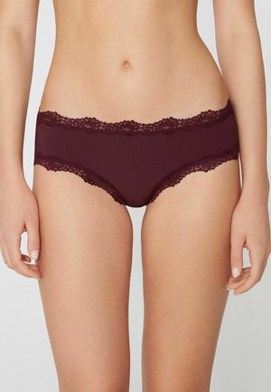 Mujer con ropa interior hipster de encaje en color rojo oscuro, mostrando el torso desde la cintura hasta la parte superior de los muslos sobre un fondo neutro.
