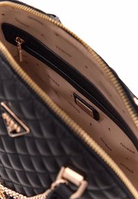 Borsa a tracolla nera trapuntata con cerniera dorata, dotata di fodera beige a motivo con logo "GUESS" e dettagli in hardware dorato.