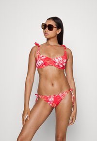 Biquíni floral vermelho com alças ajustáveis e acabamento texturizado. Óculos de sol com uma grande armação retangular castanha complementam o visual.