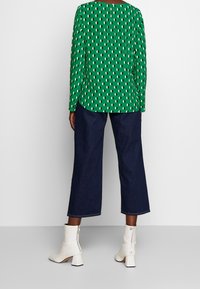 Grünes Langarmshirt mit weißem Ovalmuster, kombiniert mit dunkelblauen, weit geschnittenen Jeanshosen und weißen Stiefeletten mit Blockabsatz und Reißverschluss.