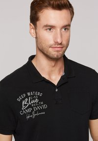 Polo nero con tessuto texture, dotato di taschino sul petto e grafiche ricamate in bianco e grigio, tra cui "Deep Waters" e "Camp David."