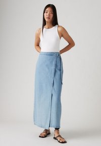 Lys blå denim wrap nederdel med en sidesløjfe, der har en lige pasform og en subtil tekstur. Sammen med en hvid ærmeløs top og sorte sandaler.