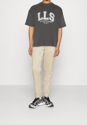 Person trägt ein dunkelgraues T-Shirt mit LLS-Logo, beige hochgekrempelte Hose und schwarz-weiße Sportschuhe und steht vor einer schlichten Wand.