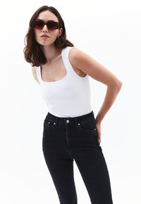 Witte mouwloze bodysuit met een vierkante halslijn, gecombineerd met hoge zwarte jeans. Model draagt schildpadbril.