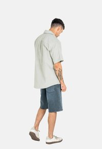 Camisa de botón de manga corta en verde claro combinada con pantalones cortos de mezclilla. Presenta un corte relajado, un yugo en la parte posterior y una textura sutil.