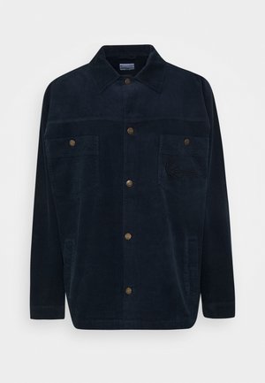 Veste-chemise en velours côtelé bleu foncé avec manches longues, fermeture à boutons sur le devant, deux poches poitrine et un logo brodé discret sur la poche gauche.