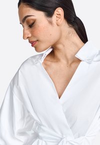 Weiße Wrap-Bluse aus leichtem Stoff, mit V-Ausschnitt, knöpfbarem Kragen und weiten Ärmeln, die an der Taille gebunden sind. Glatte Textur.