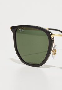 Ochelari de soare cu ramă neagră și lentile verzi, design unghiular, accente aurii și pad-uri nazale transparente, având un logo Ray-Ban pe una dintre lentile.