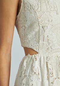 Abito in pizzo di un leggero beige con ricami elaborati e un taglio in vita. Tessuto testurizzato con dettagli arricciati all'orlo.