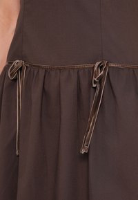 Καφέ φόρεμα με σούρα στη φούστα, διακοσμημένο με καφέ σατέν κορδόνια στη μέση και ομαλή υφή υφάσματος.