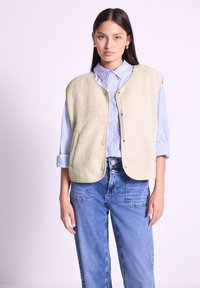 Gilet en polaire vert clair avec des bords arrondis, des boutons sur le devant et des poches latérales, porté sur une chemise bleue à col et associé à un jean bleu.