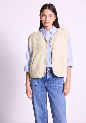 Gilet en polaire vert clair avec des bords arrondis, des boutons sur le devant et des poches latérales, porté sur une chemise bleue à col et associé à un jean bleu.