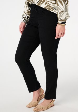 Vrouw in zwarte slim-fit broek, beige geweven slingback schoenen en een langemouwen top met een bruin en wit loofpatroon.
