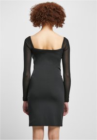 Robe noire ajustée avec de longues manches transparentes et un décolleté carré au dos, dotée d'une texture lisse et d'un ourlet au niveau des genoux.
