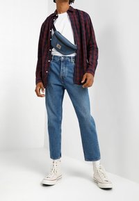 Mies, joka pukeutuu sinisiin farkkuihin, valkoisiin lenkkareihin, valkoiseen t-paitaan, auki olevaan punaisiin ja mustiin ruutupaitaan sekä siniseen olkalaukkuun yksinkertaista valkoista taustaa vasten.