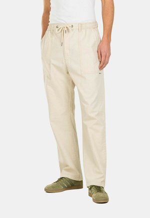 Beige cotton cargo pants with an adjustable drawstring waistband, side pockets, and a relaxed fit, paired with green sneakers.