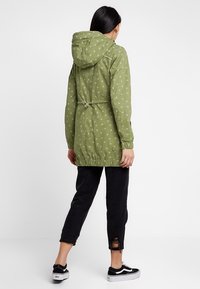 Olive green water-resistant jacket with white polka dots, adjustable hood, elastic cinch waist, and long sleeves. Styled with black pants.