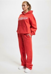 Rød hættetrøje med hvidt logo, frontlomme, parret med matchende røde sweatpants. Stoffet har en blød tekstur, komplementeret af hvide sneakers.
