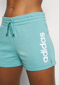 Pantaloni corti turchesi chiari realizzati in tessuto morbido, con una vita elastica dotata di cordino e un logo "adidas" bianco sul lato.