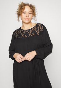 Vestido preto com uma sobreposição de renda no decote, mangas longas e um detalhe de cintura com laço. Tecido suave com um corte solto.