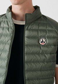 Groen, doorgestikt gewatteerd vest met een hoge kraag en ritssluiting aan de voorkant. Heeft een ronde logopatch op de linkerborst en een gladde textuur.