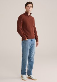 Rostfarbener Strickpullover mit einem Viertelreißverschluss, kombiniert mit hellblauen geraden Jeans und grünen Sneakers mit braunen Akzenten.