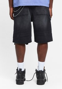 Shorts din denim negru cu o croială dreaptă, având accente din lanț pe buzunarele din spate și o textură decolorată. Purtați cu șosete albe și bocanci.
