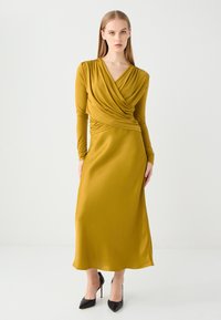 Lang mosterdkleurige jurk met lange mouwen, gedrapeerde bodice en een gladde textuur. Heeft een aansluitende taille en een vloeiende rok, gecombineerd met zwarte hakken.